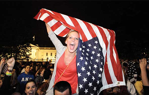 A young woman smiling, chanting, and waving an American flag in front of the White House with a big crowd behind her.