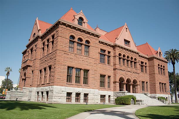 A photo showing the big three stories courthouse building with brick-like looking walls. The base level is white while the top two stories in light brown color with orange brick roof.