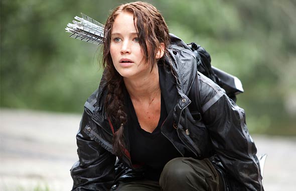 Actress Jennifer Lawrence in a scene of her portrayal of Katniss Everdeen where her messed up hair is tied up at the back of her head, she has bloody scratch wounds on her neck and chest, she wore a black vinyl jacket, black shirt, grey pants, with arrows in arrow holder on her back while she is in semi-squatting position in the woods.