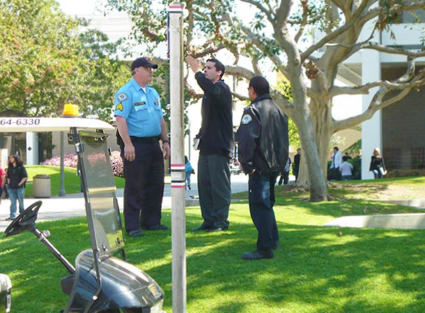 Non-student Ronald Cardiel, dressed in black pants and long sleeves shirt, standing between two campus security officers on a lawn area by the quad.