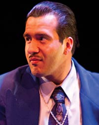 Danny Gonzalez portrait as Henry with his hair combed back and wearing blue jacket and tie with with white shirt and gold chain with cross pendant.