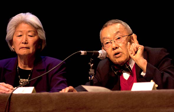 Dr. Donal Teruo Hata sharing testimonies in front of a microphone while Hatsuko Mary Higuchi watches on next to him.