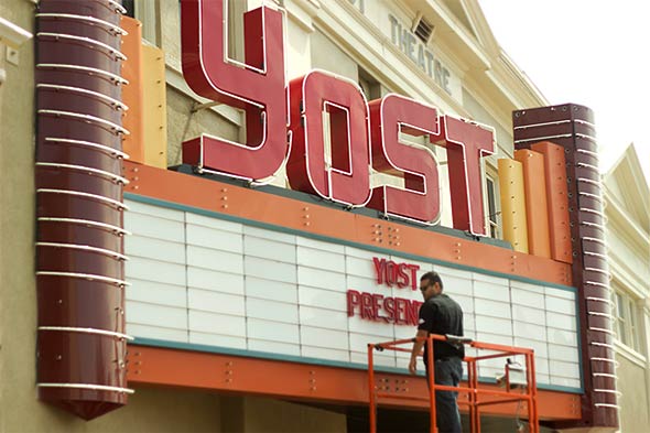 A worker on top of construction stand in front of big and colorful YOST sign outside Yost Theater placing sign which reads "YOST PRESENT…"