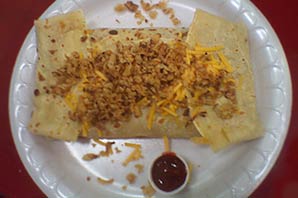 A golden crepe folded like a burrito on a plate with nutty and cheese toppings and a side of sauce.