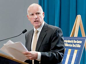 Governor Jerry Brown sitting at a podium with speaker and hands in his paper and some statical charts displayed behind him.