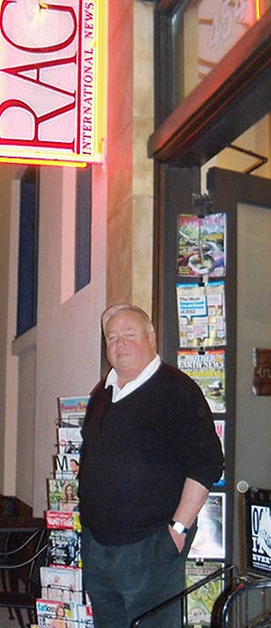 James Kendrick, owner of RAG-International-News-Stand, wearing a white shirt with dark color sweater stand in front of his store with rags of magazines and newspapers by the side of the store.