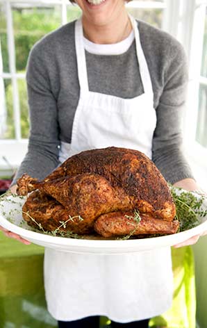 A woman carrying a full sized roasted turkey, with the top part of the woman's head cut off the photo, only showing her smile.